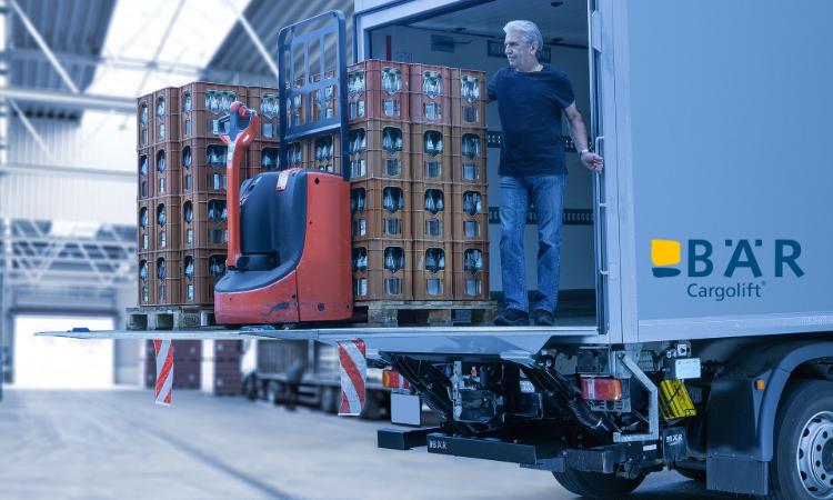Řidič stojí s několika paletami s nápoji a elektrickým paletovým vozíkem na plošině zadní hydraulické zdvihací plošiny BAR.